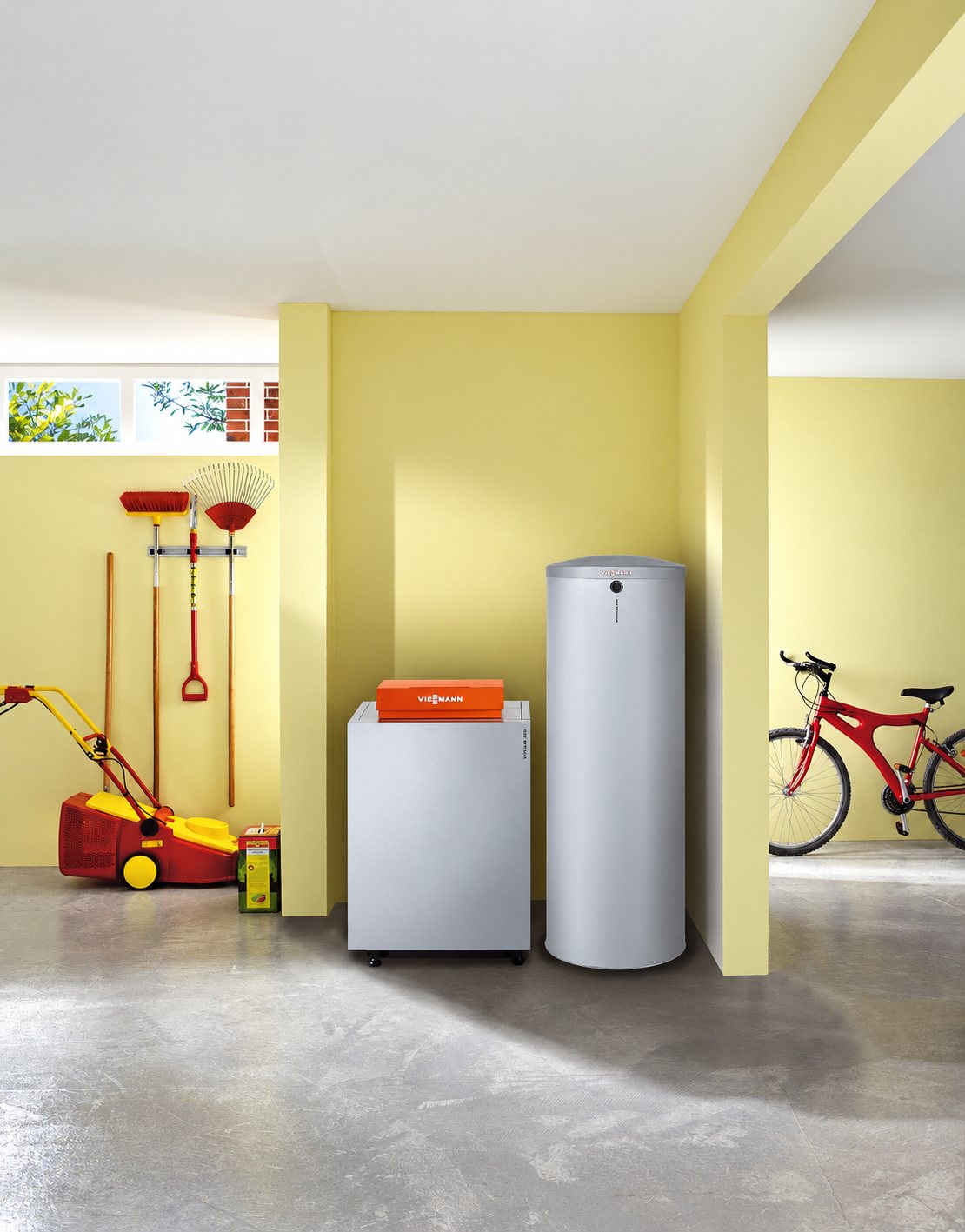 Une chaudière grise surmontée d'un boîtier orange et un grand ballon d'eau chaude sont installés dans un garage aux murs jaunes, avec des outils de jardin, une tondeuse et un vélo rouge.