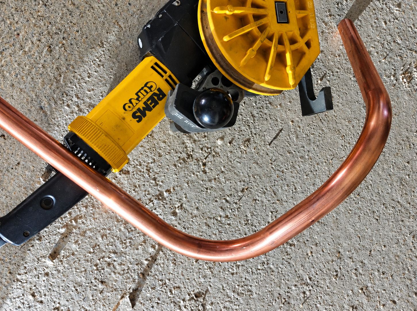 Une cintreuse électrique jaune REMS Curvo plie un tube de cuivre brillant contre un mur en béton brut.