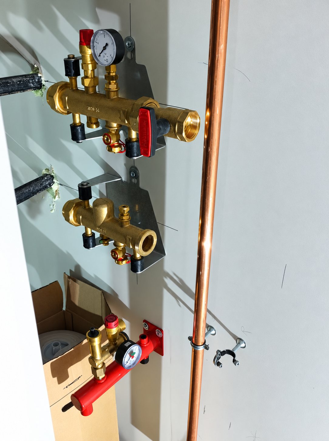 Des collecteurs de plomberie en laiton avec manomètres sont fixés au mur d'un local technique, à côté d'un tuyau de cuivre vertical.