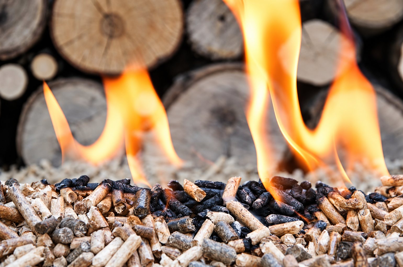 Gros plan de granulés de bois en train de brûler avec de hautes flammes orange, devant une pile de bûches floues.