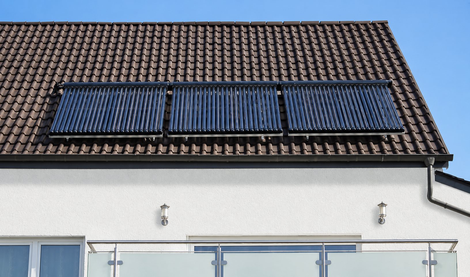 Trois capteurs solaires thermiques à tubes sous vide installés sur le toit de tuiles sombres d'une maison blanche avec balcon en verre.