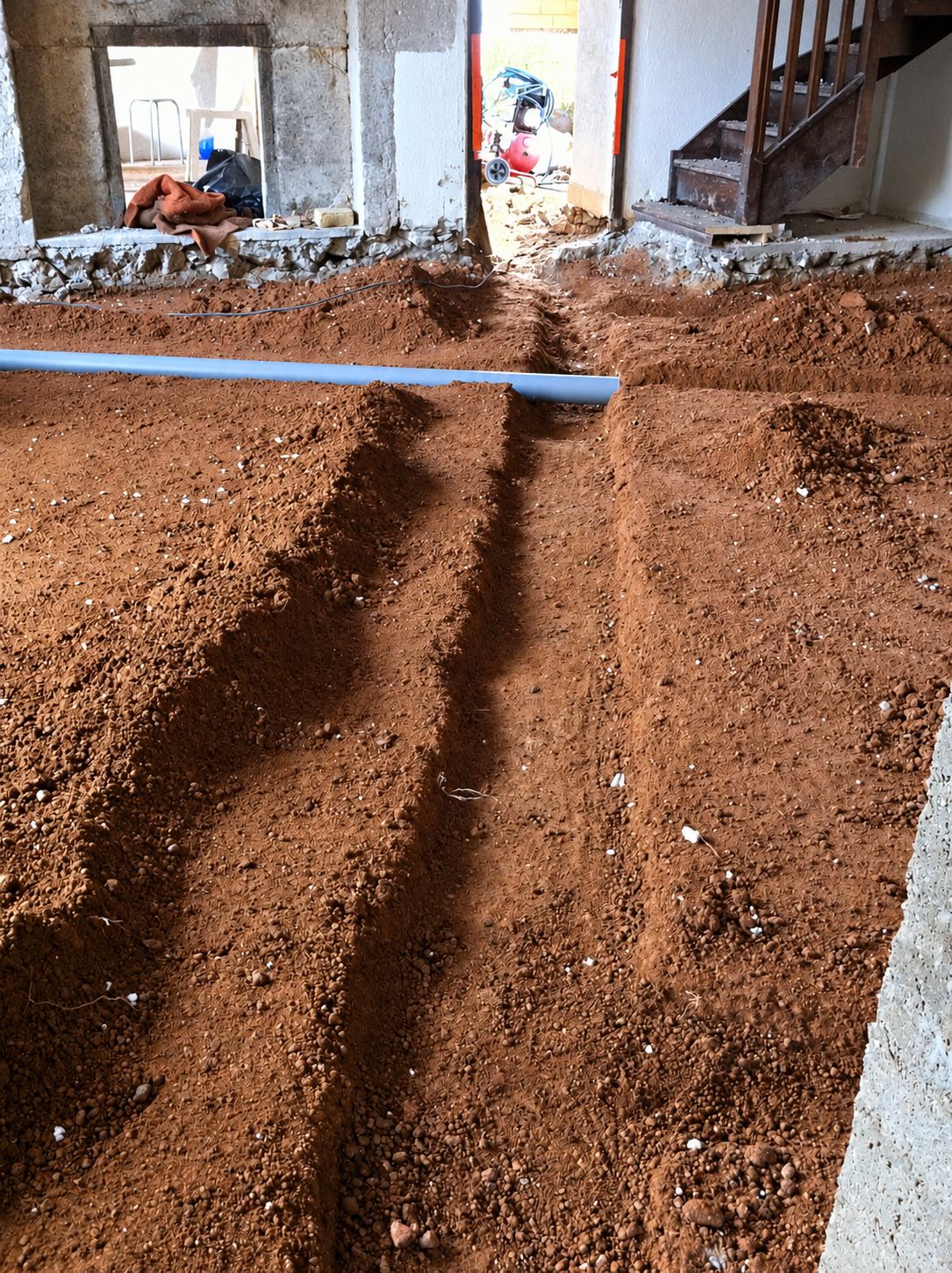 Intérieur d'une maison en travaux dont le sol en terre est creusé de tranchées, avec un tuyau bleu posé en travers et un escalier en bois au fond.