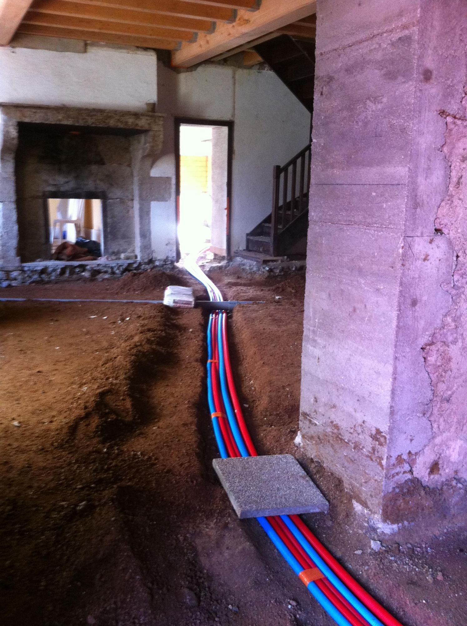 Dans une pièce en rénovation au sol de terre, des tuyaux rouges et bleus sont posés dans une tranchée près d'un pilier, avec une cheminée en pierre et un escalier en bois au fond.