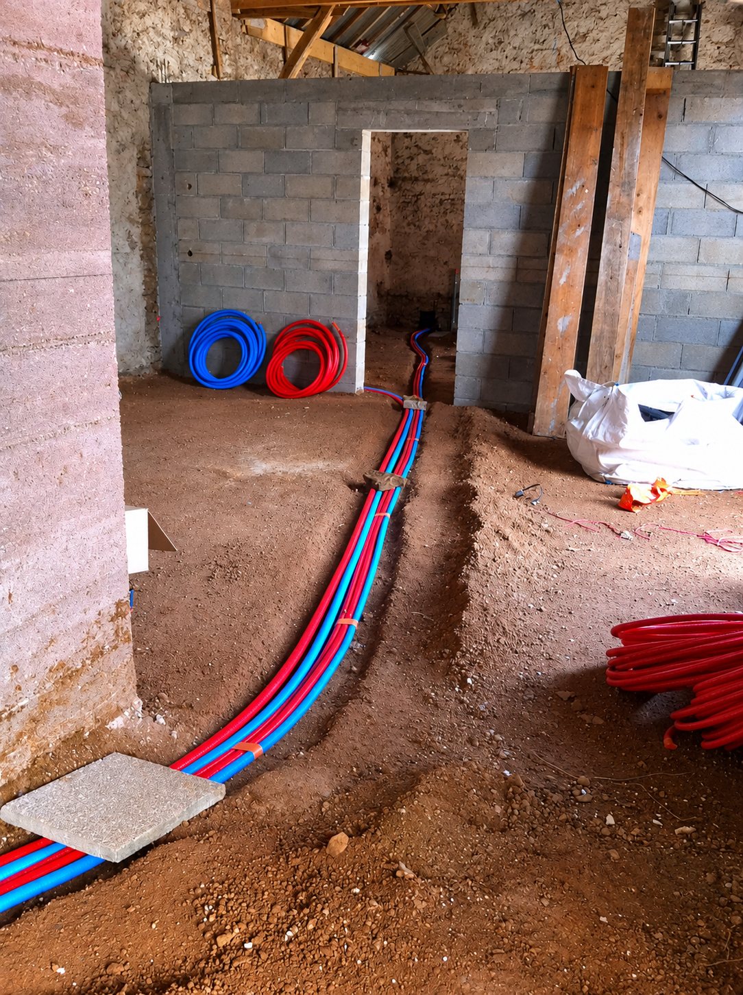 Dans un bâtiment en rénovation, des tuyaux de plomberie rouges et bleus sont posés dans une tranchée creusée dans la terre, près de murs en parpaings.